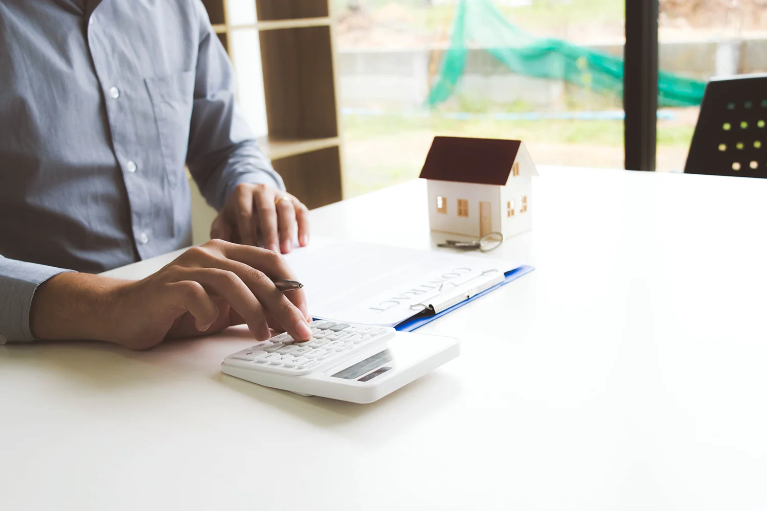A person sitting at a desk with a calculator, papers, and a small house model, preparing to sign a contract or manage financial calculations.