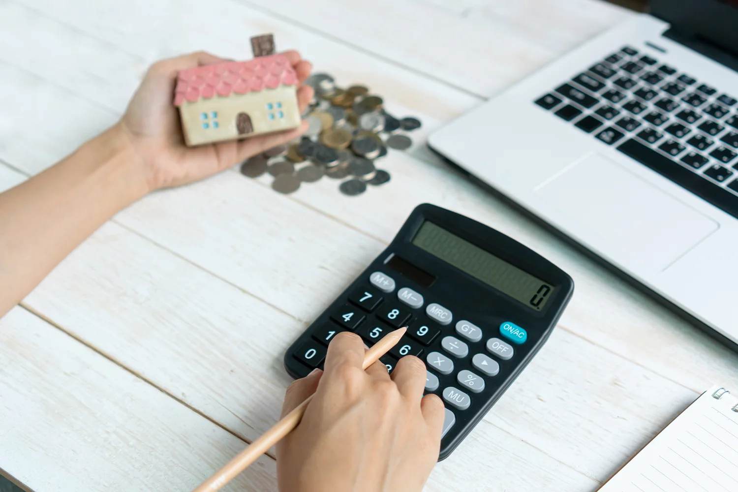 Hand holding a house model while using a calculator, with coins scattered on a table, symbolizing property tax calculations and savings through Tax Cutter’s services.