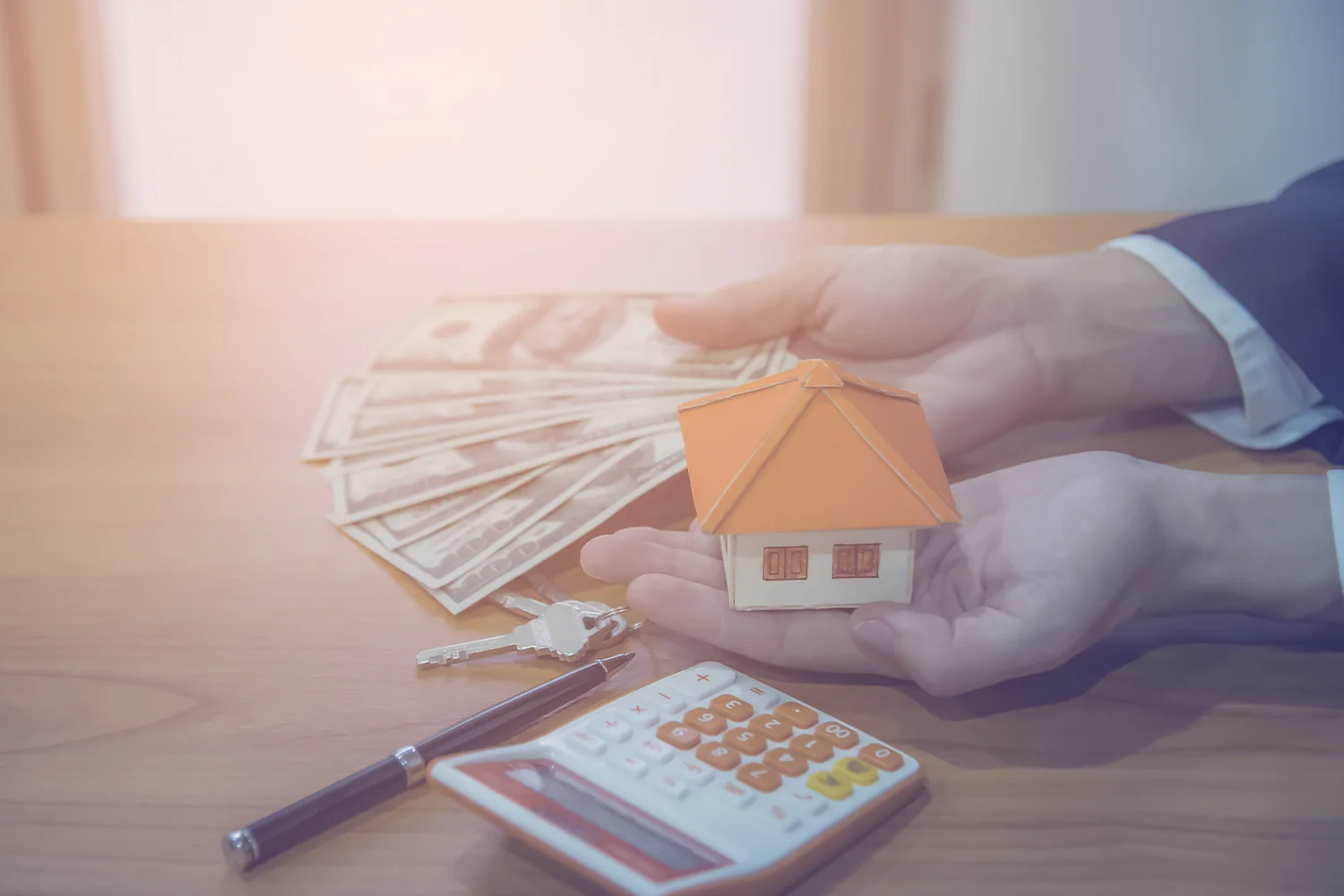 Hands holding a miniature house and money, symbolizing savings from property tax protests, with a calculator and keys on the table for tax reduction services by Tax Cutter.
