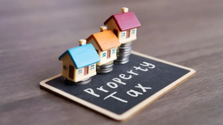 Three miniature houses placed on stacks of coins over a board labeled "Property Tax," representing rising property taxes and home value costs.