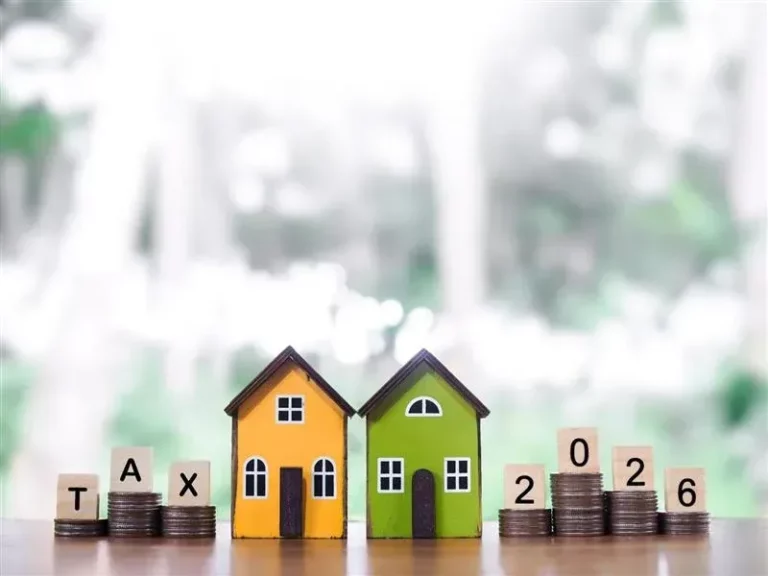 Two miniature houses with stacks of coins and wooden blocks spelling “TAX 2026,” representing property taxes and financial planning.