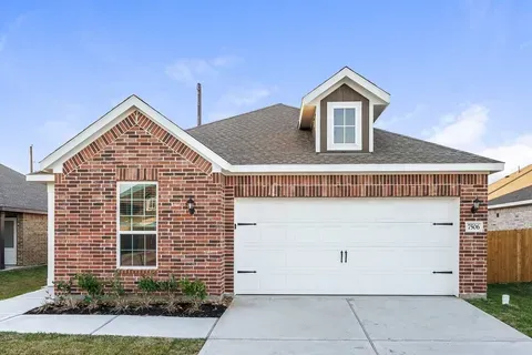 Single-story brick suburban house with a white garage door, pitched roof, front window, and small landscaped yard under a clear blue sky.