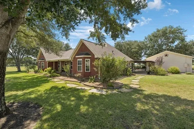 Single-story brick home with a landscaped yard, surrounding trees, and a detached structure connected by a walkway under a clear blue sky.