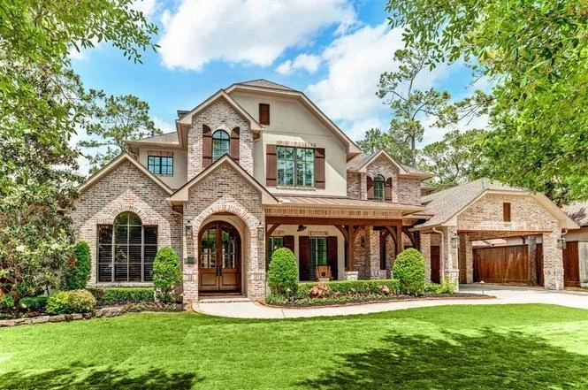 Large two-story brick home with arched windows, a covered front porch, wooden garage doors, and a landscaped lawn surrounded by trees.