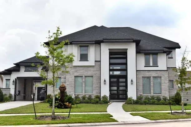 Modern two-story house with a stone and stucco exterior, large front door, landscaped yard, and driveway in a suburban neighborhood.