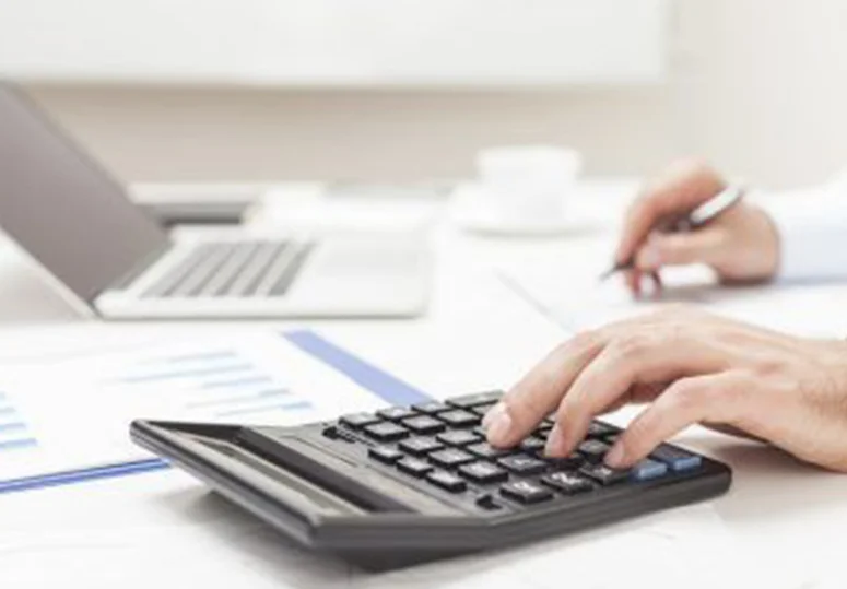Person using a calculator with financial charts and laptop on desk, representing tax calculations, property assessment, and financial planning.