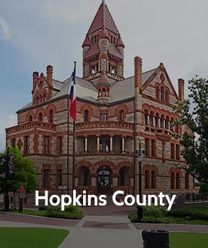 Hopkins County Courthouse in Texas with red brick exterior, detailed arches, and a central clock tower in a landscaped square.