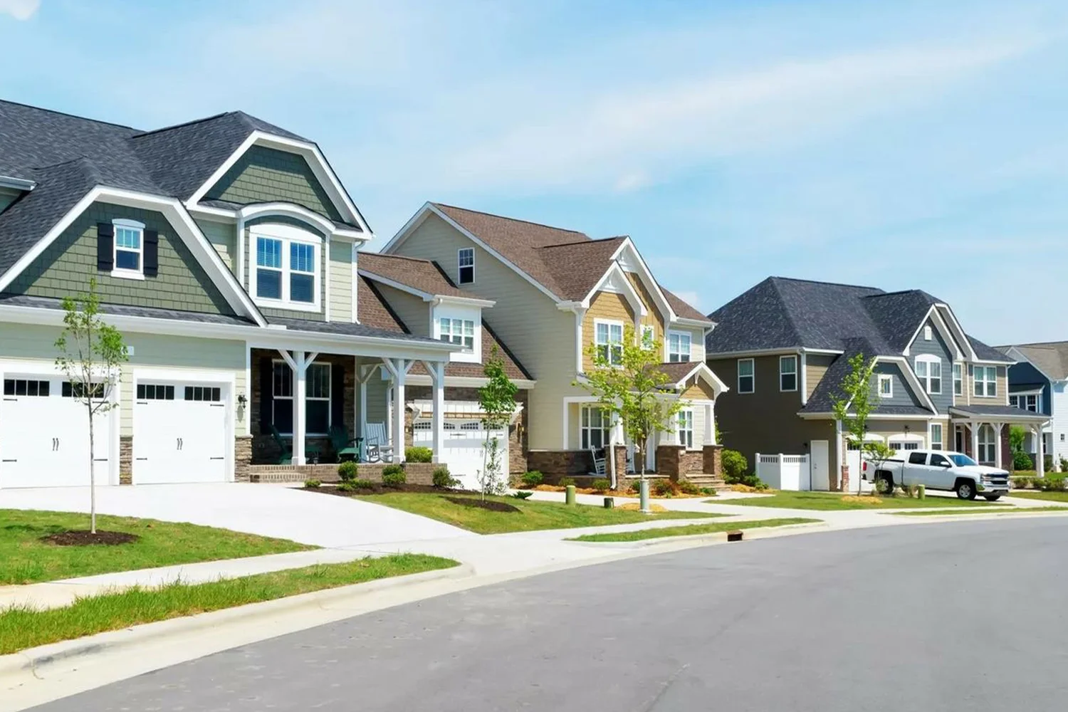 Row of modern suburban homes with garages, driveways, and landscaped lawns along a quiet residential street.