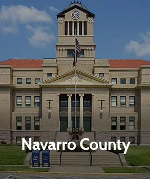 Navarro County Courthouse with a tall central clock tower, red roof, and symmetrical stone façade under a clear blue sky.