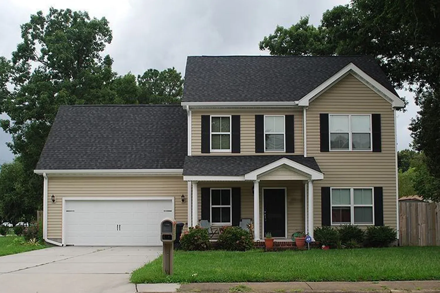 Two-story suburban house with beige siding, black shutters, attached garage, and front lawn in a residential neighborhood.