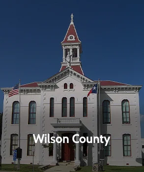 Wilson County Courthouse in Texas with white facade, red-roofed clock tower, and American and Texas flags at the entrance.